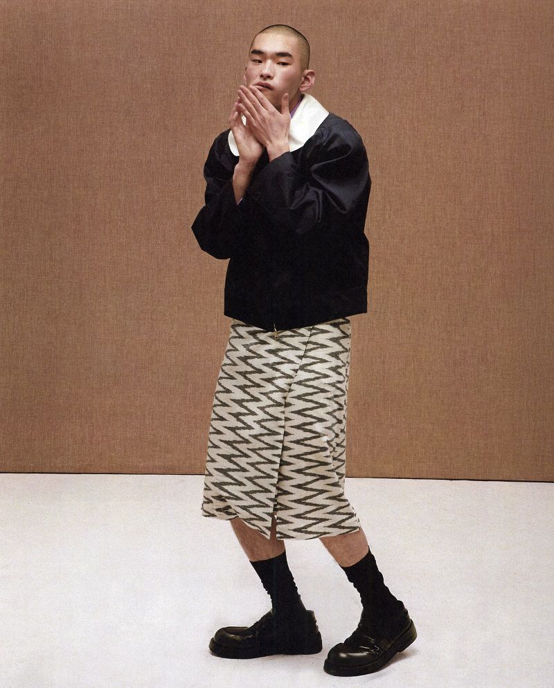 Man with shaved head wearing a black jacket and patterned skirt against a textured studio background.