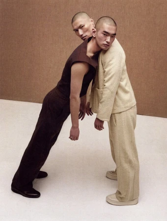 Two East Asian male models with buzz cuts pose together in brown and cream menswear against a warm neutral backdrop.