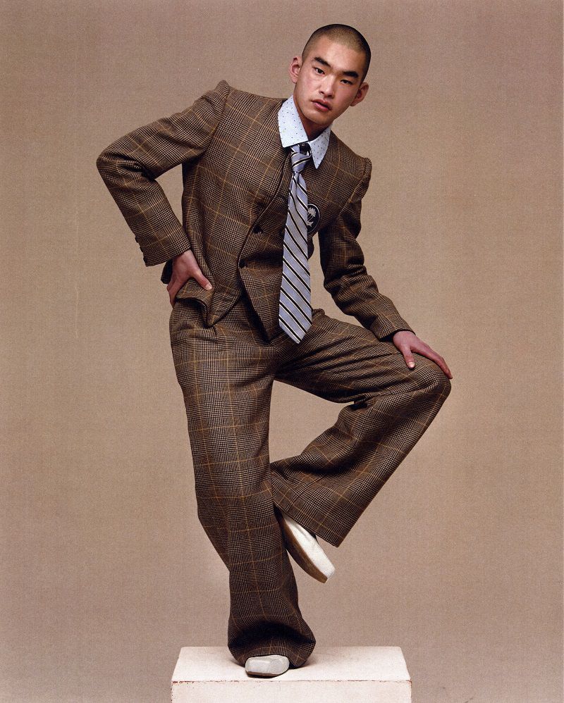 East Asian man in a brown plaid suit, collared shirt, and striped tie balancing on one leg on a pedestal against a beige backdrop.