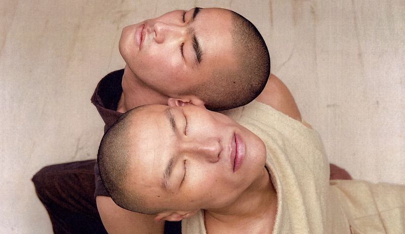 Two East Asian models with shaved heads resting their heads together with closed eyes against a neutral backdrop.