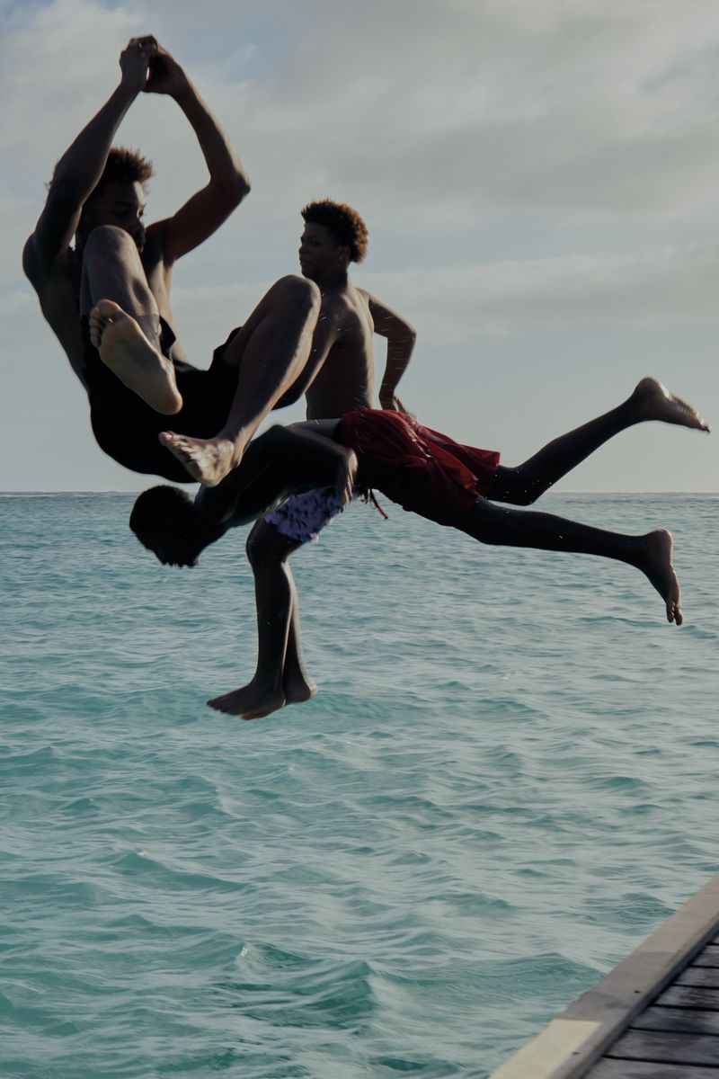 Three young men jumping and flipping into clear turquoise ocean water from a wooden dock.