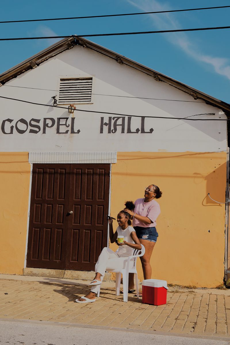 Two Black women outside a yellow Gospel Hall building, one seated on a white plastic chair styling the other standing behind her