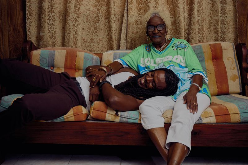 An elderly Black woman in a green jersey sits on a sofa as a Black man rests his head on her lap in a warmly lit room.