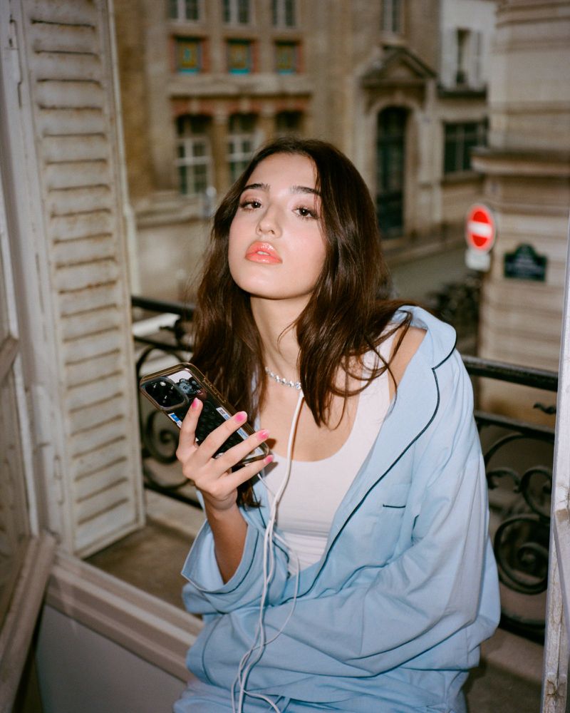 Léonie Cassel seated by a Parisian window in a light blue pajama top holding a smartphone with wired earphones.