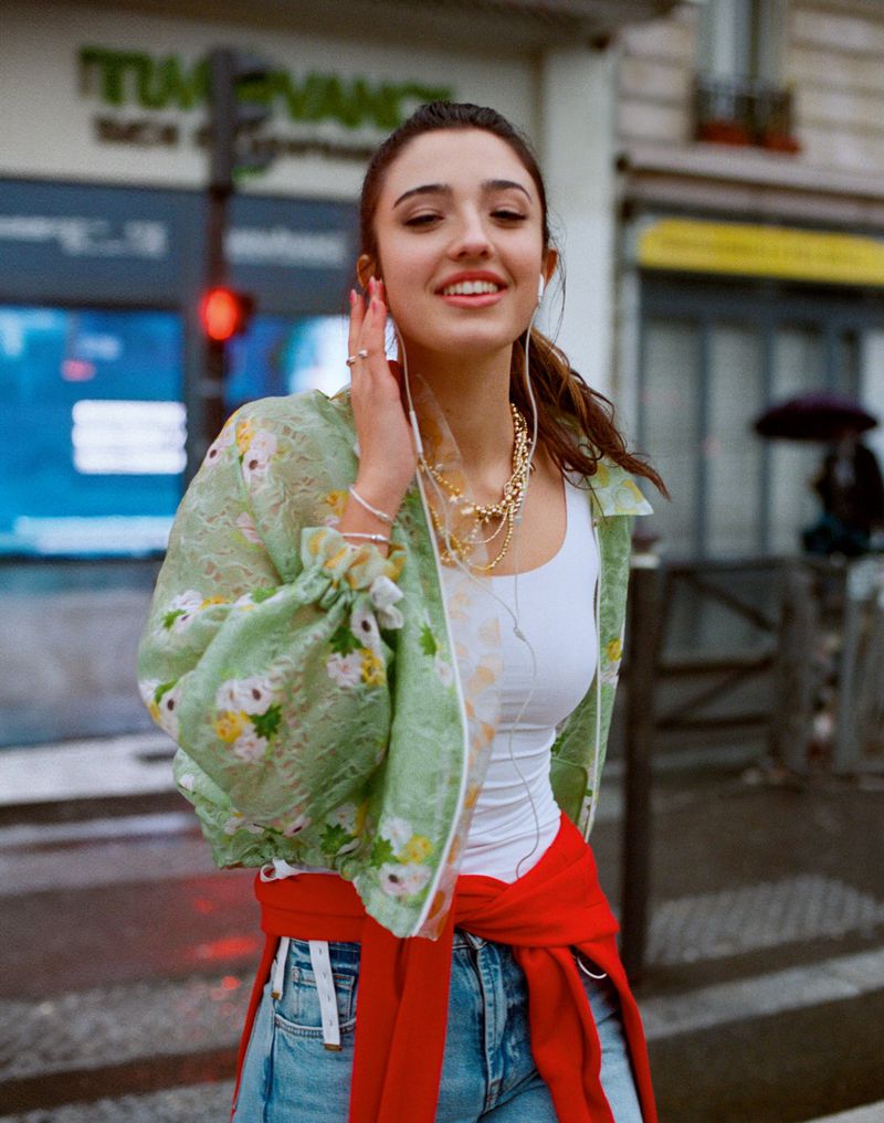 Léonie Cassel smiling in a sheer floral green jacket, white tank top, and red sweater tied at her waist on a city street.
