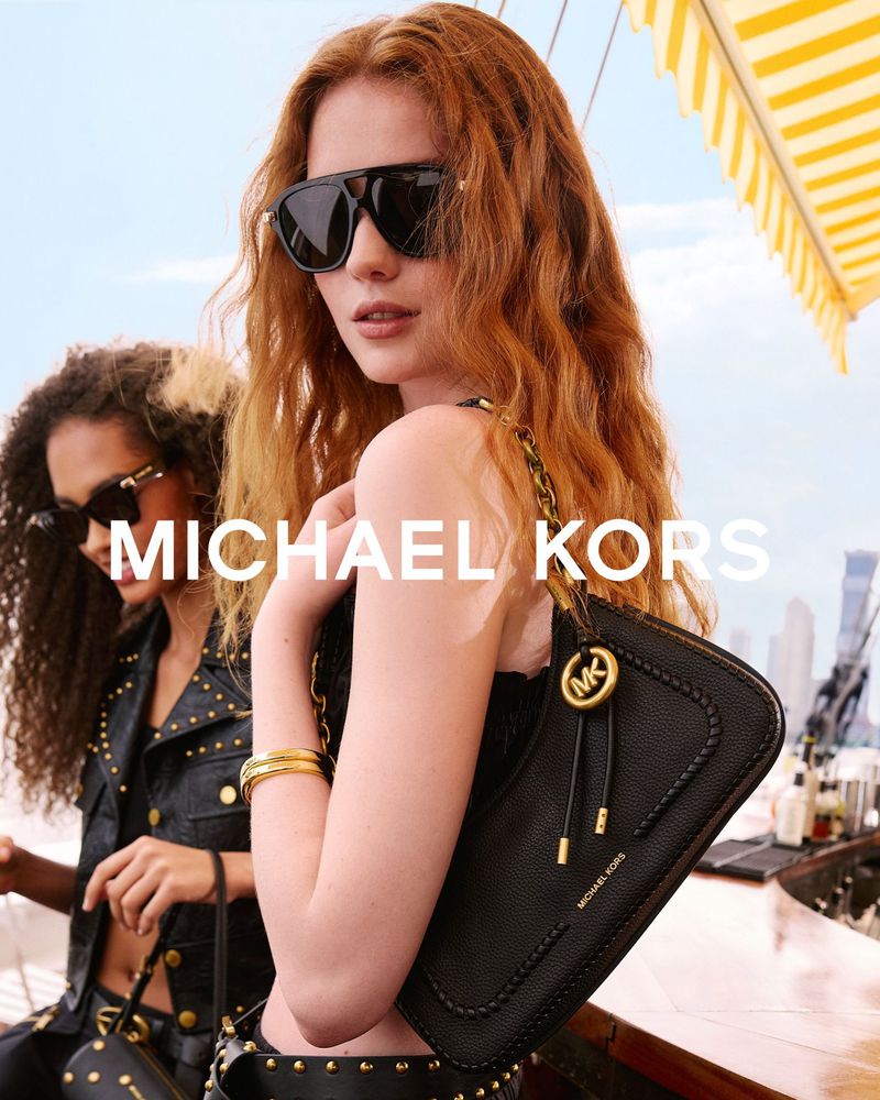Redhead model in black sunglasses holding a black leather Michael Kors shoulder bag with gold chain detailing outdoors.