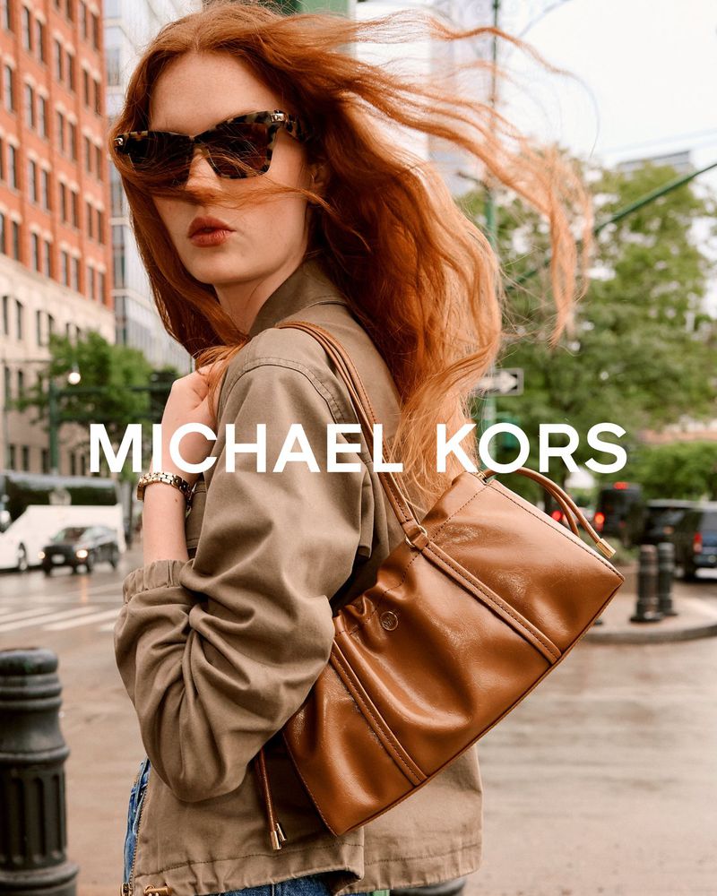 Woman with windblown red hair wearing sunglasses and an olive jacket carrying a tan leather shoulder bag on an urban city street.