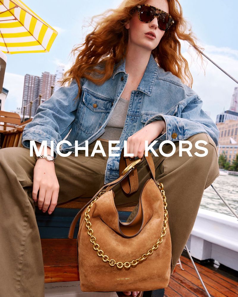 Model with long red hair wearing a denim jacket and holding a brown suede shoulder bag on a boat