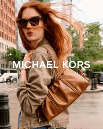 Woman with windblown red hair wearing sunglasses and an olive jacket carrying a tan leather shoulder bag on an urban city street.