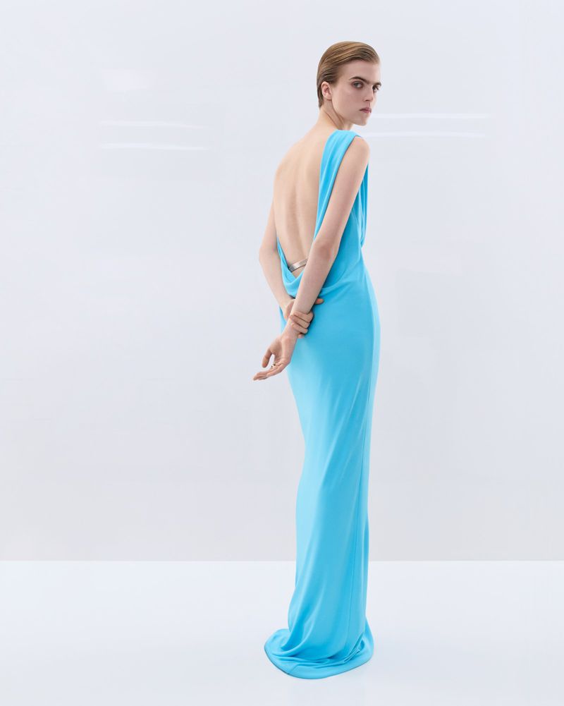 Model in a long vibrant sky blue backless gown looking over her shoulder against a seamless white background.