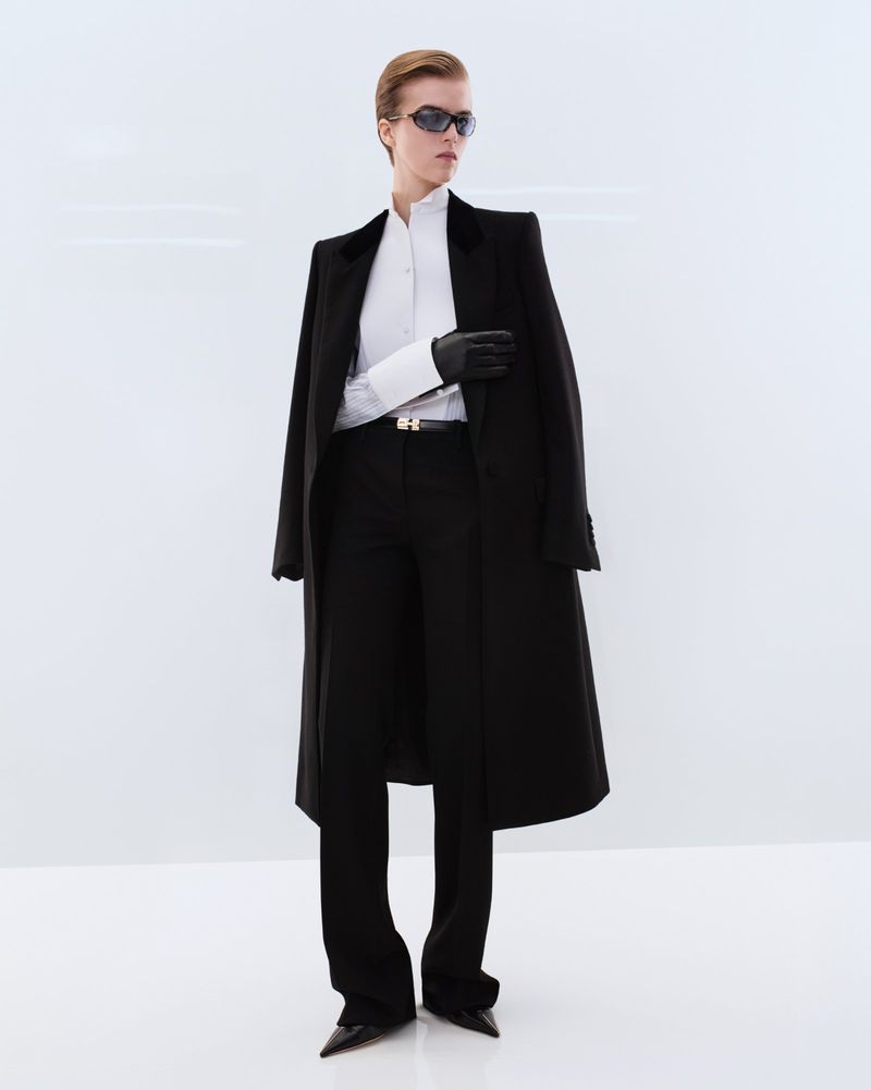 Model wearing dark sunglasses and a long black tailored overcoat over a white shirt against a white seamless background.