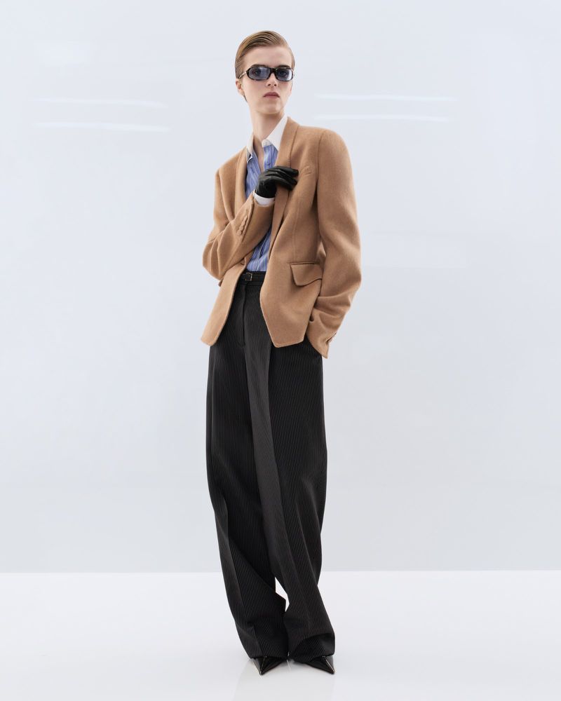 Model wearing dark sunglasses, a tailored camel blazer, and wide-leg black trousers against a white seamless studio backdrop.