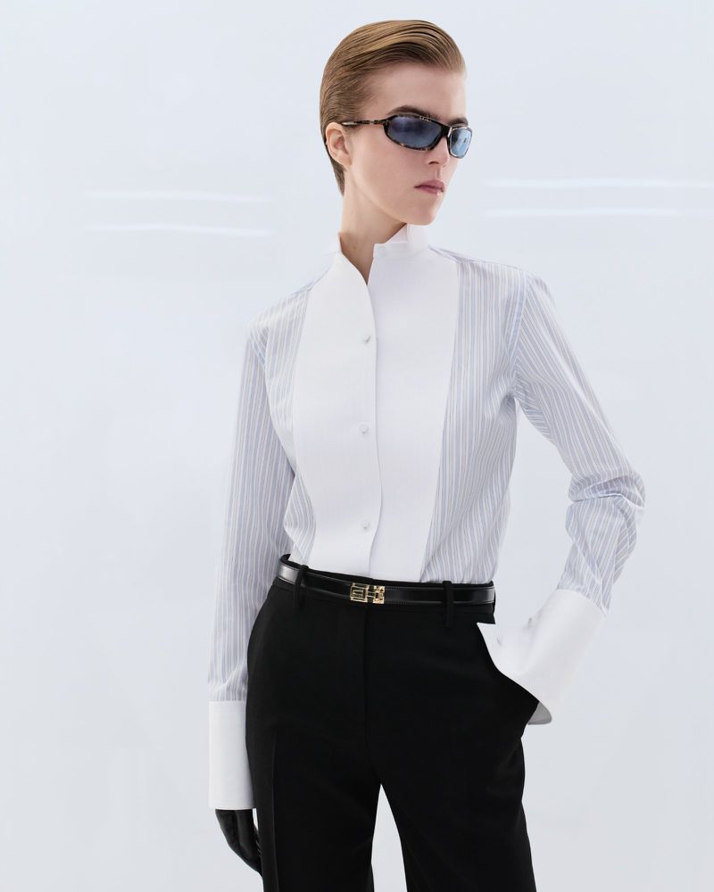Model wearing dark sunglasses, striped shirt with white bib, and black trousers against a neutral background