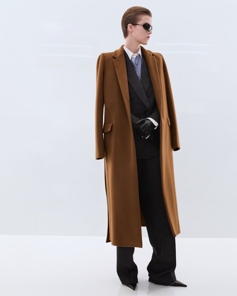 Model wearing a long camel wool overcoat, dark tailored suit, sunglasses, and leather gloves against a white studio background.