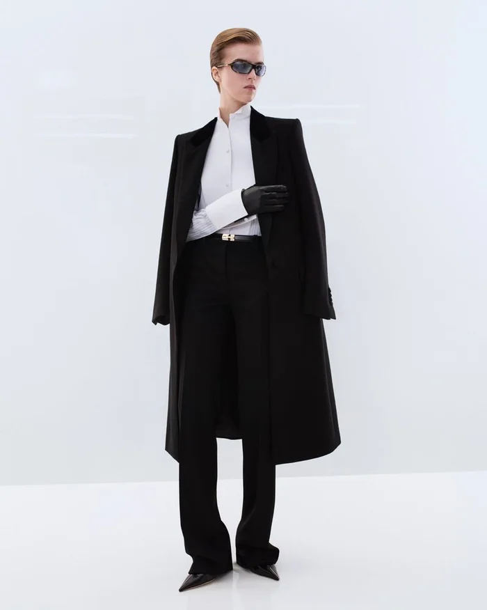 Model wearing dark sunglasses and a long black tailored overcoat over a white shirt against a white seamless background.