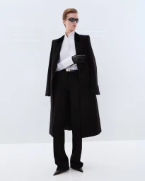 Model wearing dark sunglasses and a long black tailored overcoat over a white shirt against a white seamless background.