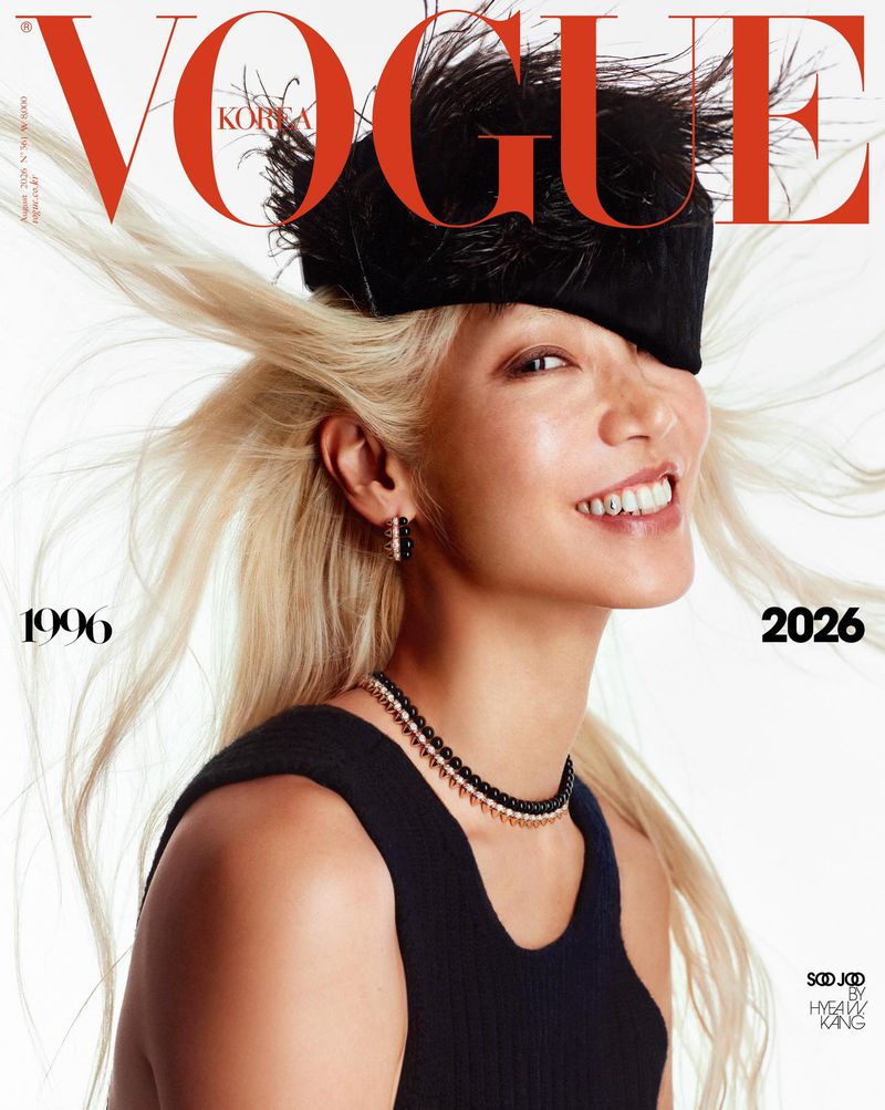 Model Soo Joo Park smiling with windblown blonde hair wearing a black feathered hat and top against a white backdrop.