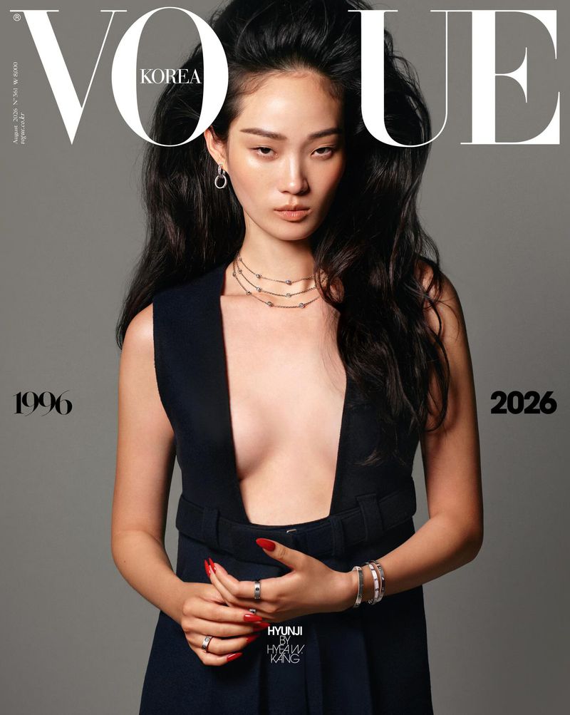 Model Hyunji Shin posing for the Vogue Korea cover wearing a deep-plunge sleeveless black belted garment against a grey backdrop.