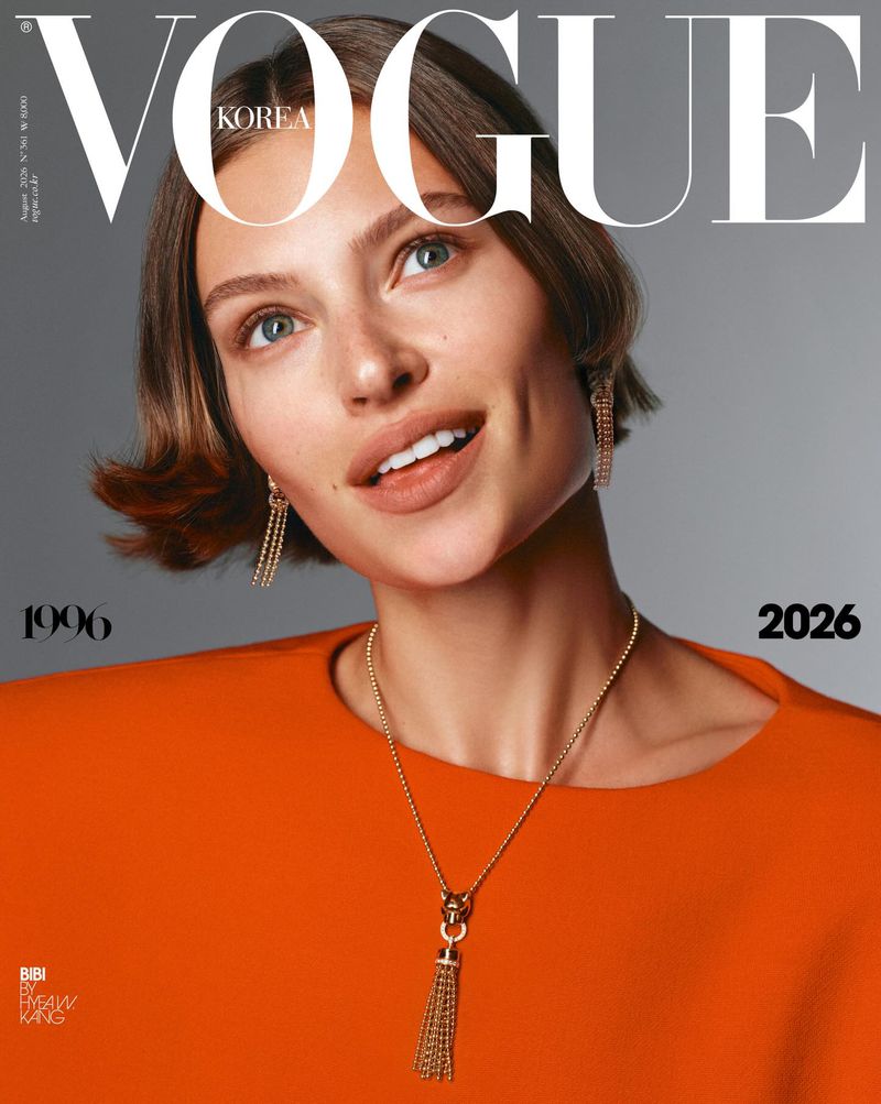 Close-up portrait of a woman wearing an orange top, gold pendant necklace, and dangling earrings against a grey studio backdrop.