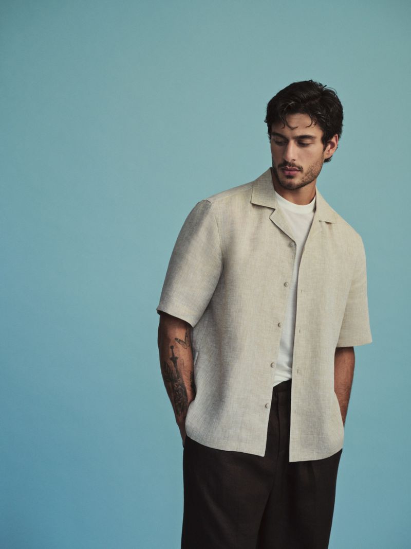 Model Ariyan Akaltun wearing a beige short-sleeve button-up shirt and dark trousers against a solid light blue studio backdrop.