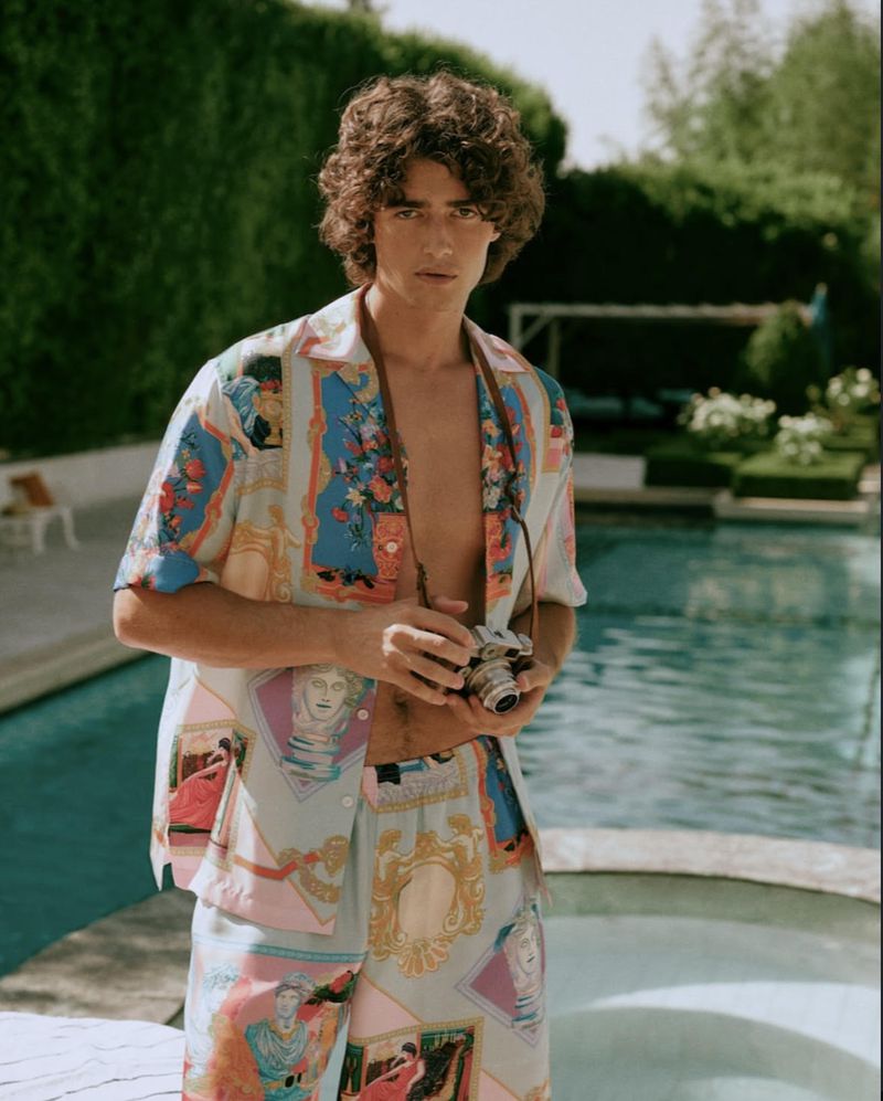 Adult male poses outdoors by pool, wearing a patterned shirt and shorts, holding a camera with green foliage background.