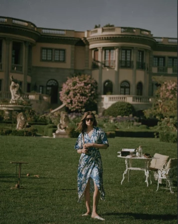 Adult female in a patterned wrap dress stands barefoot on green grass in front of a luxury home with a table and lawn game.