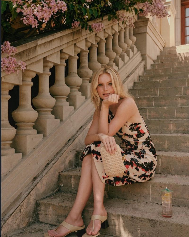 Elegant woman in floral dress seated on stone steps near pink blossoms, holding clutch, with a drink beside her in outdoor setting.