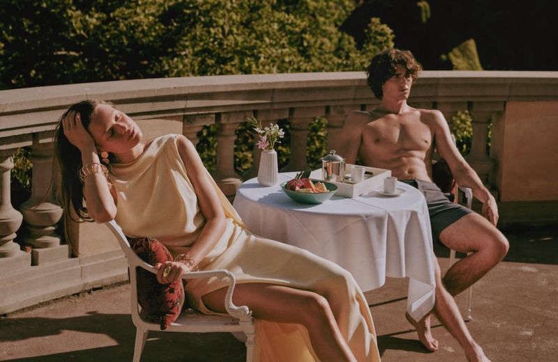 A couple sits on a sunlit balcony near a table with fruit. The woman wears a pale yellow dress, and the man is shirtless.