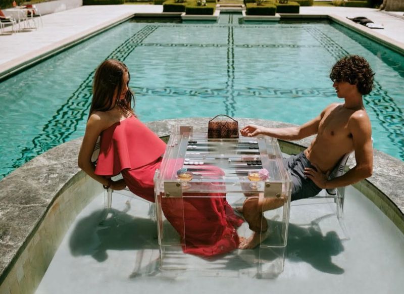 A couple plays backgammon at a transparent table in a pool. The woman wears a red dress, the man is shirtless, and there's a purse on the table.