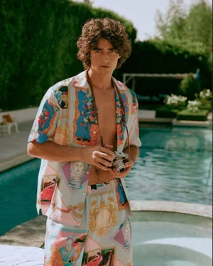Young man with curly hair wearing a printed silk shirt and shorts, holding a vintage camera by a swimming pool.