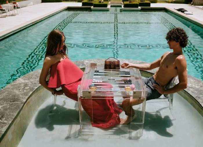 A woman in a red dress and a shirtless man playing backgammon on a clear acrylic table by a swimming pool for Neiman Marcus