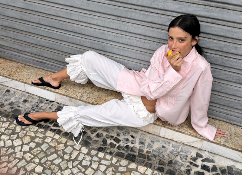 Livia Nunes Marques reclining on a curb in a pink shirt and white ruffled trousers while biting into a yellow fruit.