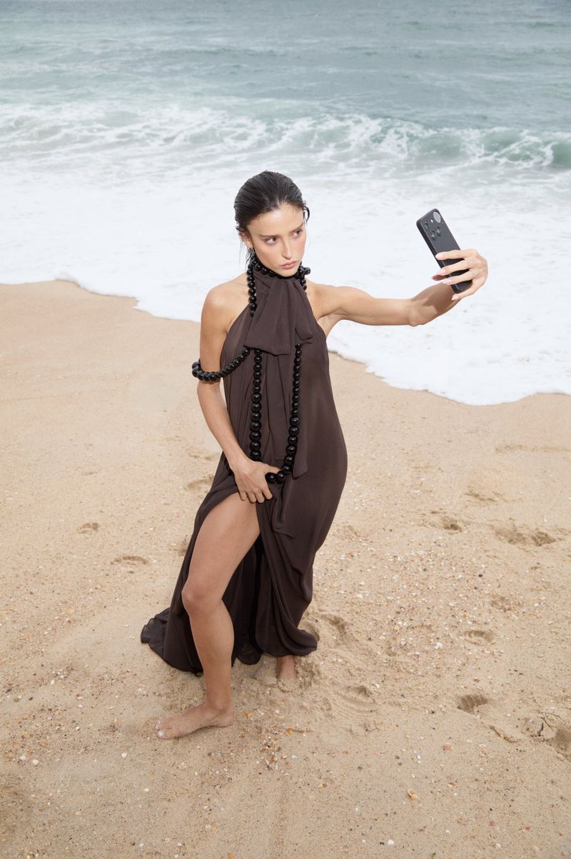 Livia Nunes Marques posing with a smartphone in a brown slit dress with black beaded rope on a sandy ocean beach.
