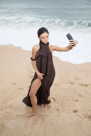 Livia Nunes Marques posing with a smartphone in a brown slit dress with black beaded rope on a sandy ocean beach.