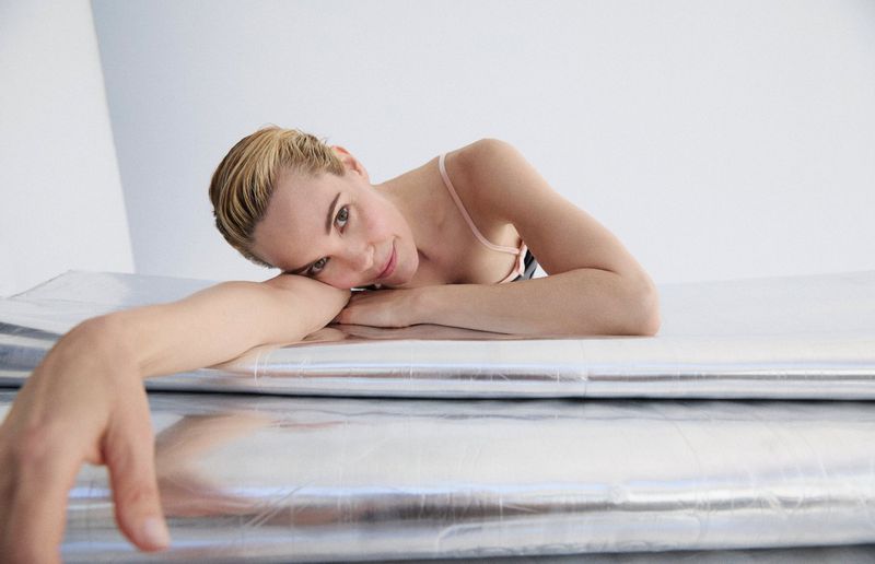 Woman with blonde hair poses in a light pink top while lying on a metallic silver surface in a brightly lit, neutral environment.