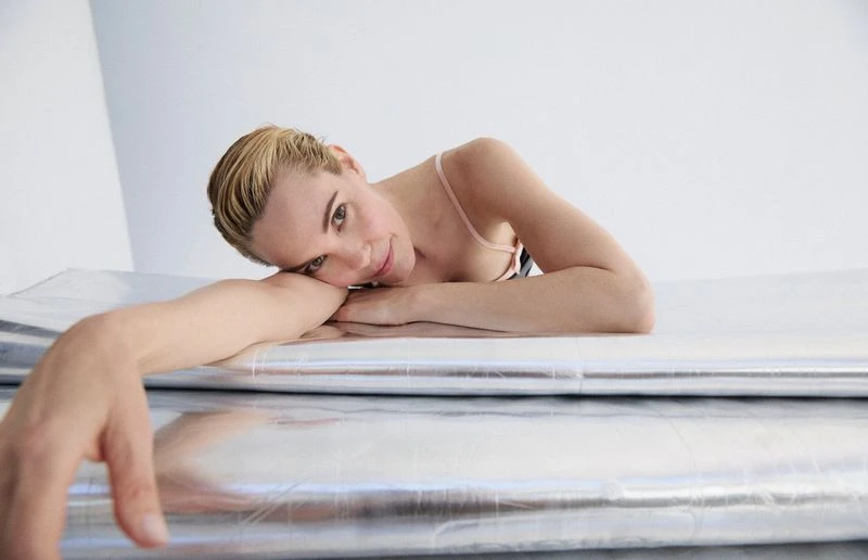 Woman with blonde hair poses in a light pink top while lying on a metallic silver surface in a brightly lit, neutral environment.