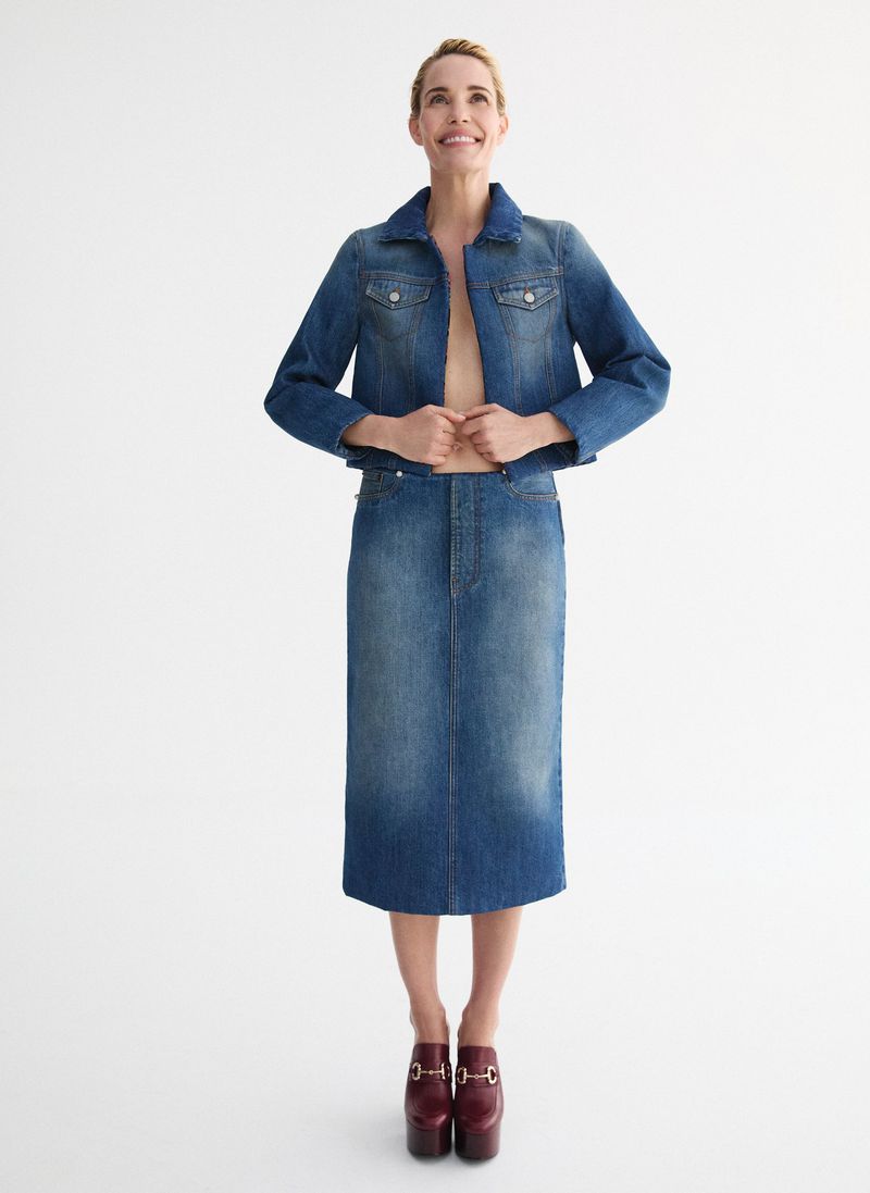 Full-length shot of a smiling woman wearing a denim jacket, denim skirt, and platform shoes, posing against a white background in a studio.