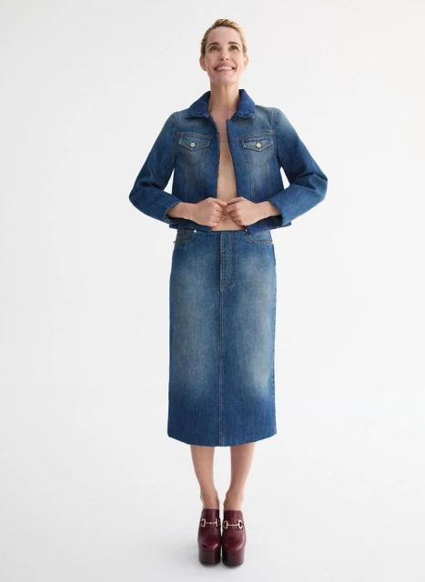 Full-length shot of a smiling woman wearing a denim jacket, denim skirt, and platform shoes, posing against a white background in a studio.