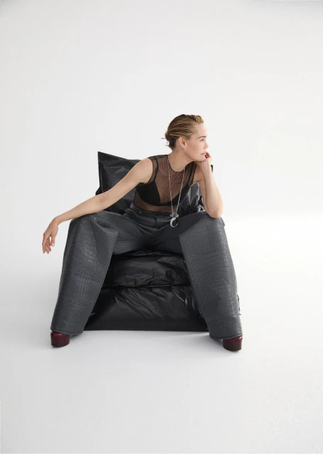 Adult woman sitting in an oversized chair, wearing a mesh top, wide leg pants, and ruby shoes, in a minimalist white photo studio.