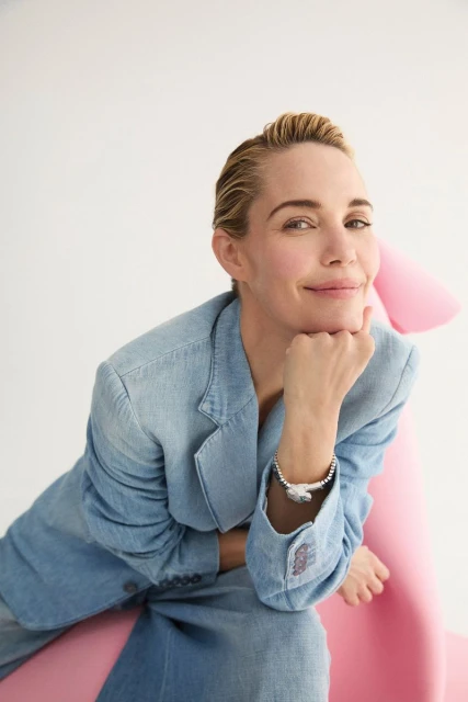 Woman in a denim blazer poses indoors with a hand on her chin, accessorized with a silver bracelet. The background is white and pink.