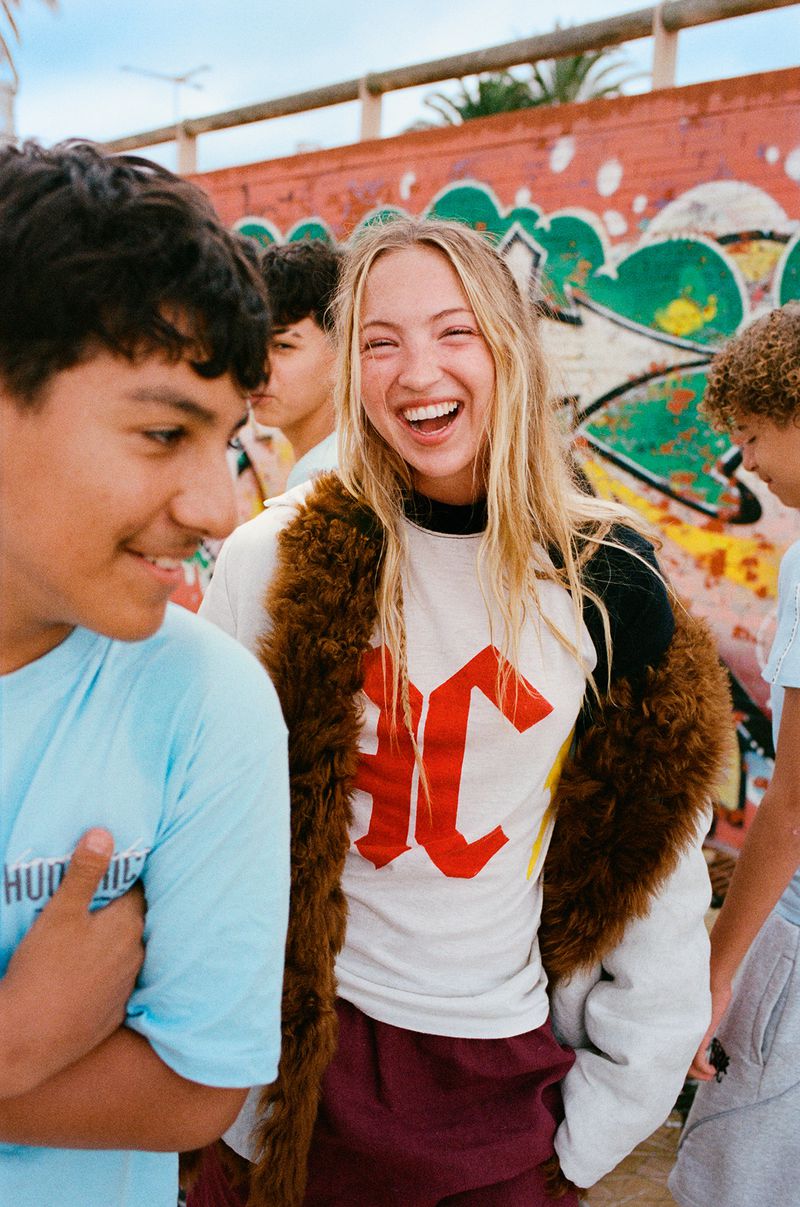 Model Lila Moss laughing outdoors wearing a graphic long-sleeve tee and faux fur jacket surrounded by teenagers near a graffiti wall.