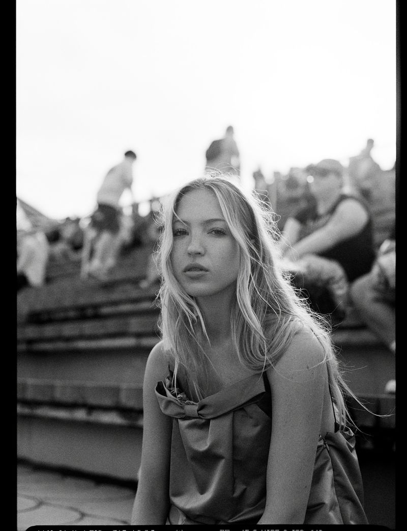 Black and white portrait of model Lila Moss wearing a draped sleeveless top while seated in front of outdoor bleachers.