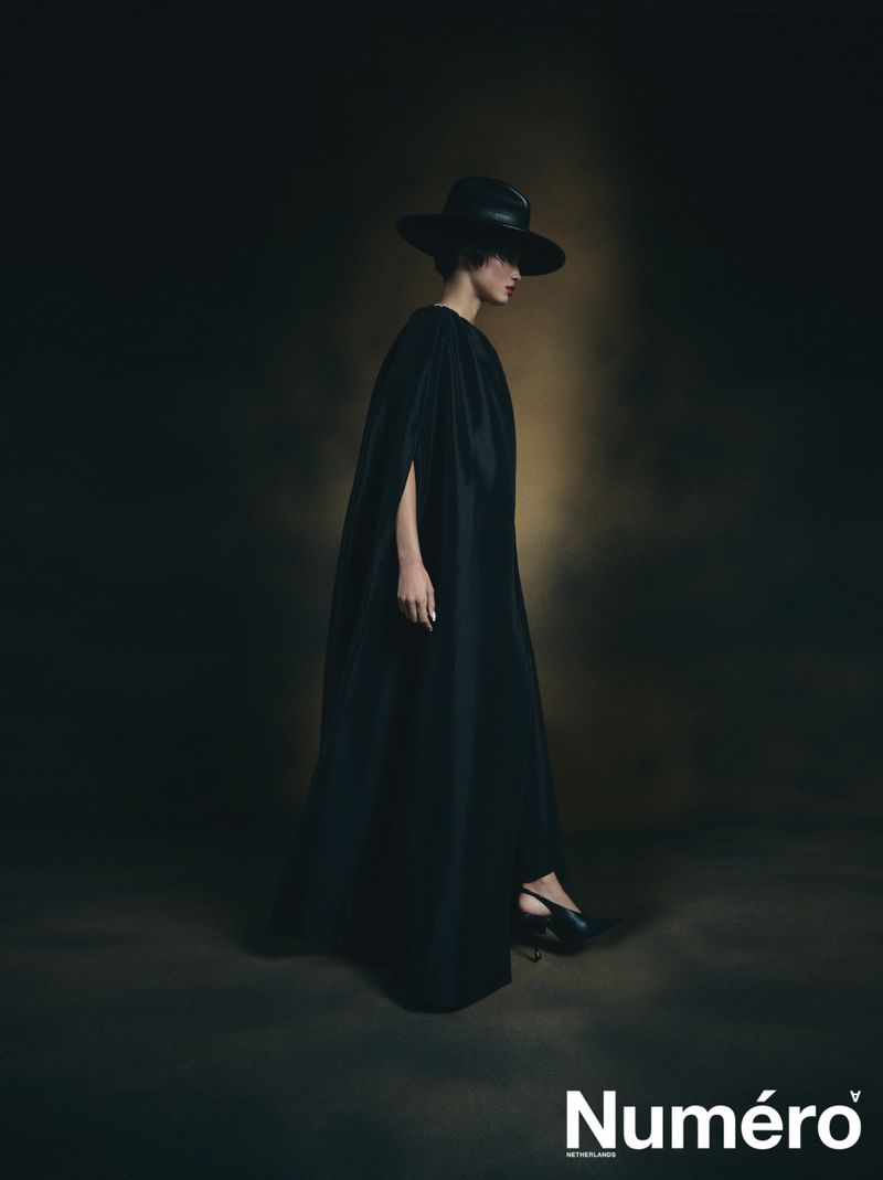 A woman in a long black cape and wide-brimmed hat poses against a dark backdrop in a fashion editorial shot for Numéro magazine.