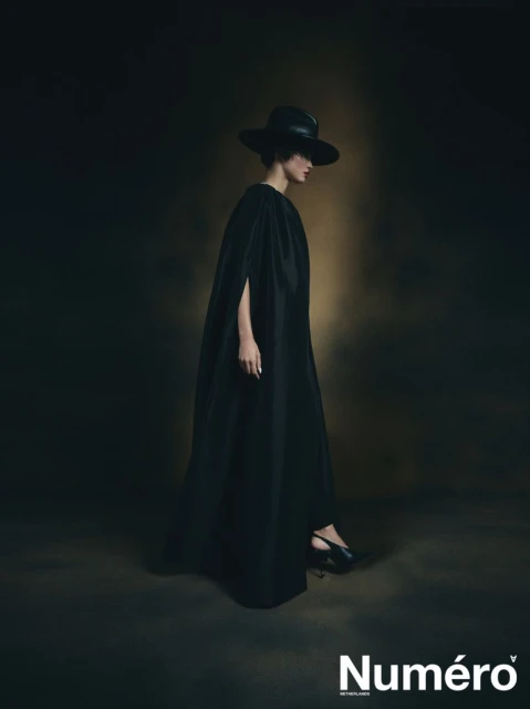 A woman in a long black cape and wide-brimmed hat poses against a dark backdrop in a fashion editorial shot for Numéro magazine.