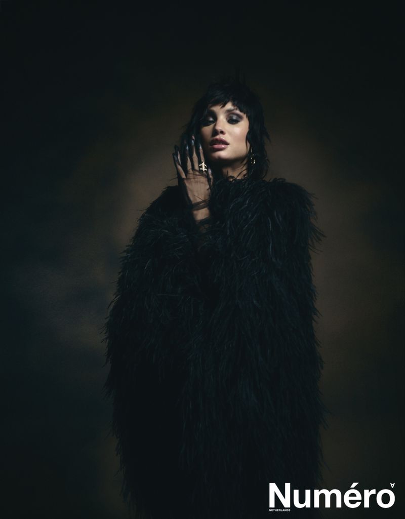 Portrait of a woman with dark hair and makeup wearing a feathery black coat in a studio with soft lighting for Numero magazine.
