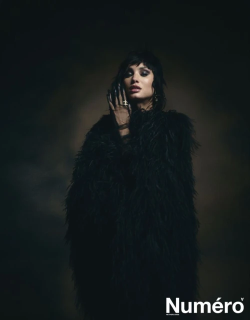 Portrait of a woman with dark hair and makeup wearing a feathery black coat in a studio with soft lighting for Numero magazine.