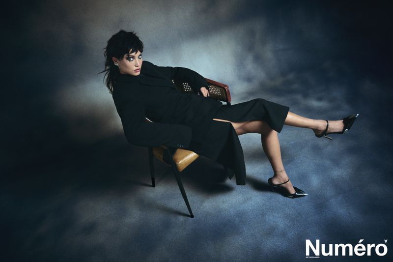 Fashion portrait of a woman in a black outfit sitting in a chair against a moody, textured backdrop, styled for a magazine layout.