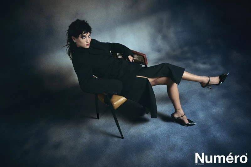 Fashion portrait of a woman in a black outfit sitting in a chair against a moody, textured backdrop, styled for a magazine layout.