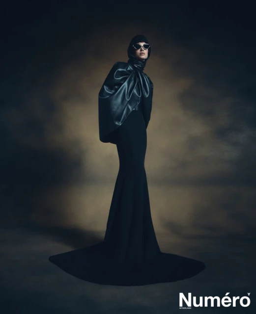 A fashion editorial shot featuring an adult woman in an elegant black dress with a large leather top and dark sunglasses posing indoors.