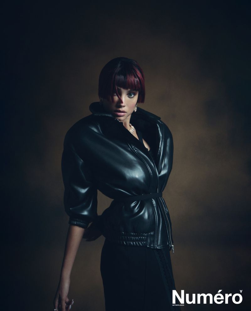 A model poses indoors wearing a black leather jacket and dark pants against a blurred brown background in a fashion editorial photo.